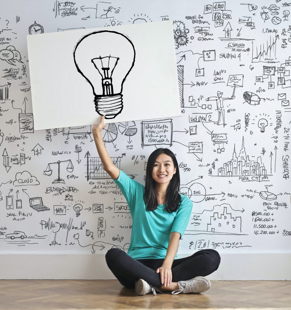 Asian woman holding up a light bulb with doodles on the wall, exploring ideas and creativity.