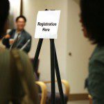 A group of people attending conferences standing in front of a sign that says registration now.