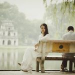 An introverted man and woman sitting awkwardly on a bench next to a pond.