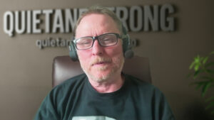 A middle-aged man with glasses and a headset sits in a chair, with the blurred text "QUIET AND STRONG" on the wall behind him, embodying introvert strengths and quiet success like David Hall.